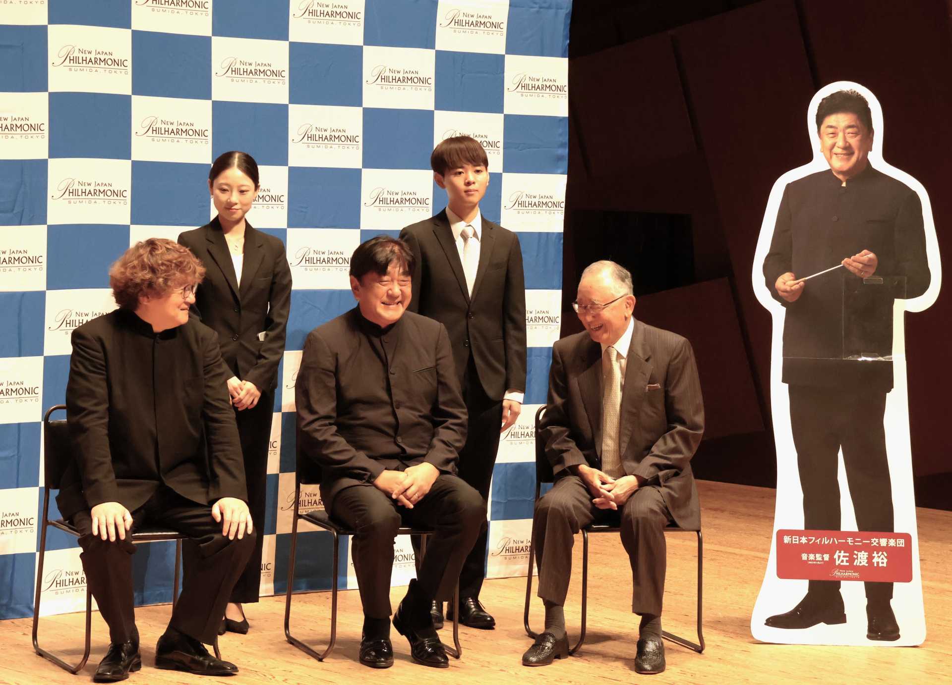佐渡裕さんの「すみだ音楽大使」のパネルの横で記念撮影（前列左から新日本フィルコンサートマスター・崔文洙氏、音楽監督・佐渡裕さん、宮内義彦理事長、後列左からＮＪＰアカデミー第１期生でクラリネット奏者・野辺かれんさん、同ホルン奏者・多田凌吾さん）