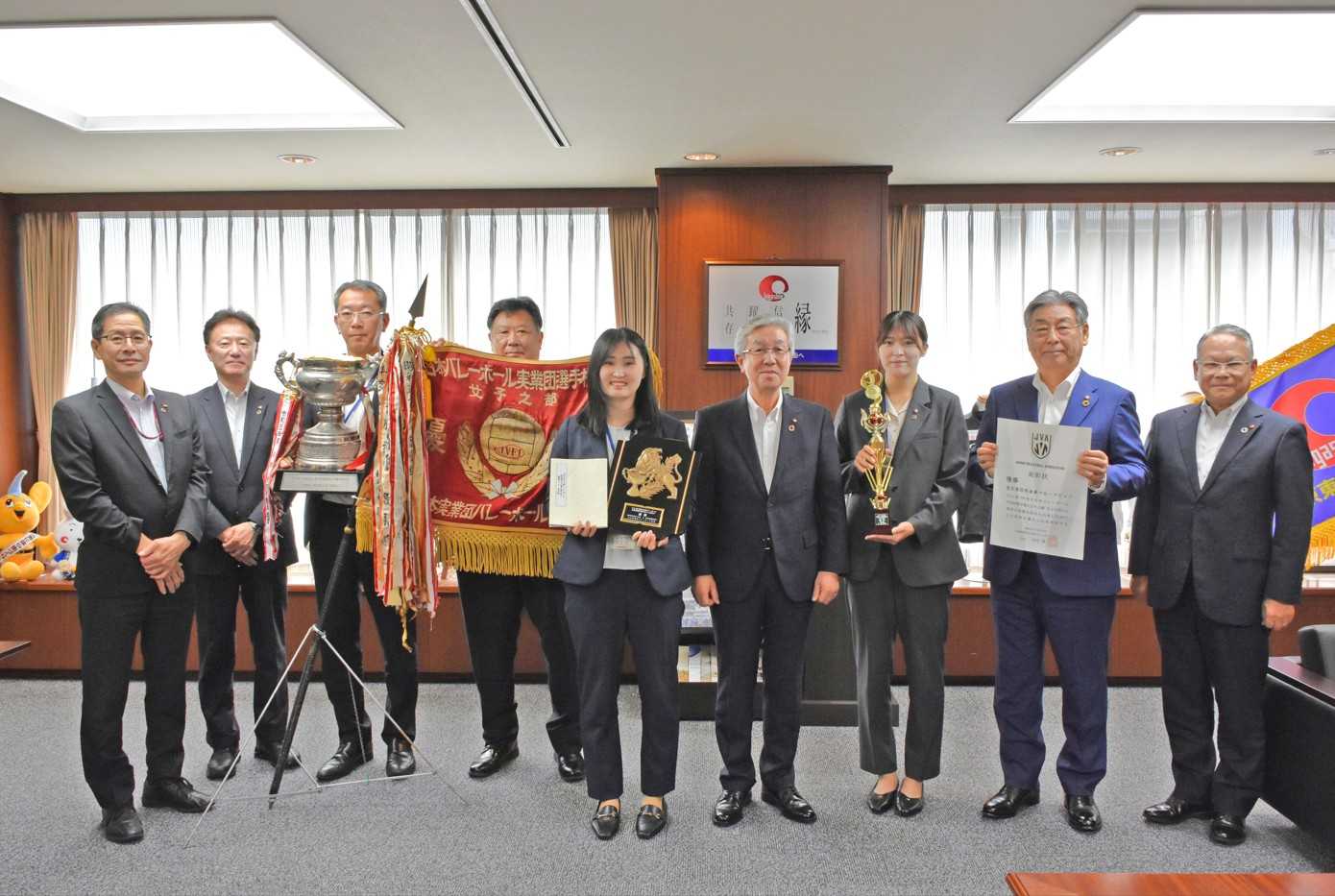 優勝報告するブルーラビッツ・大図有美監督（中央）と加藤愛菜主将（右から３人目）を称える東京東信用金庫の中田清史会長（同４人目）、吉田誠理事長（同２人目）、三石智章副理事長（右）、鈴木幹夫専務理事（左から２人目）、ブルーラビッツ顧問の金田隆夫常勤理事（左）。優勝杯を持つブルーラビッツの藤村重次部長、優勝旗を掲げる地域支援部・大橋誠司部長